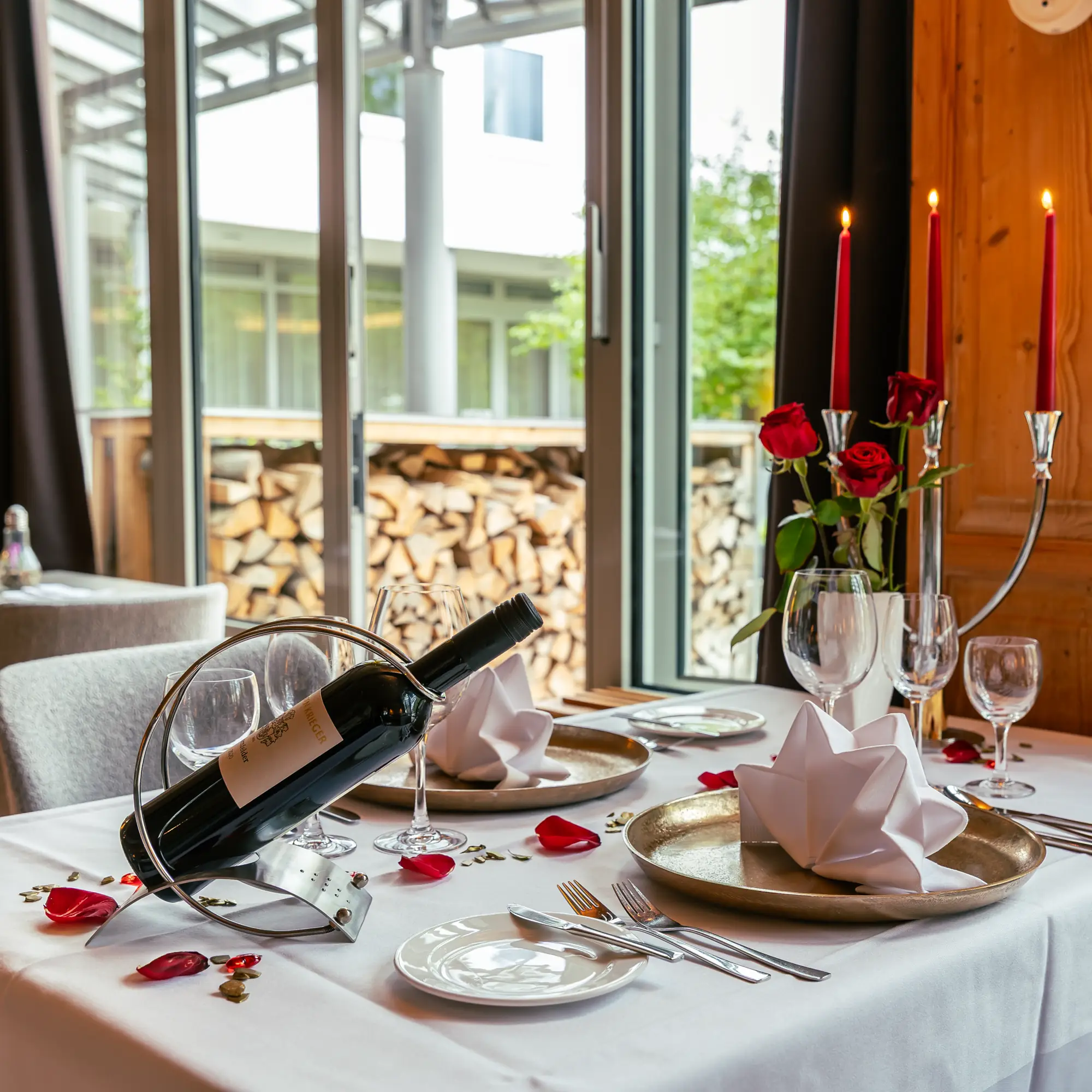 Eleganter gedeckter Tisch mit weißem Tischtuch, Weinflasche in Halter, roten Rosen, Kerzen und kunstvoll gefalteten Servietten in einem Restaurant mit Blick nach draußen.