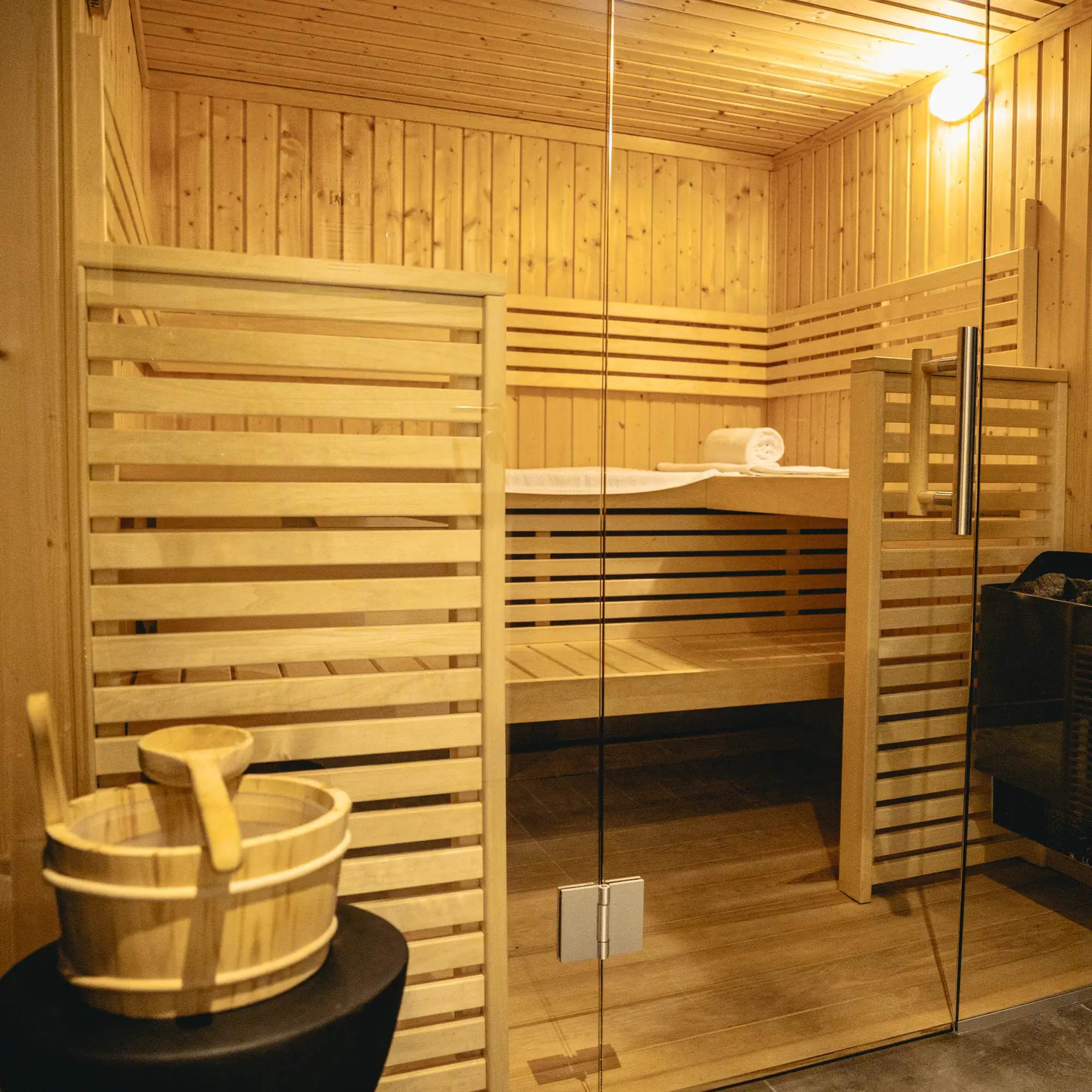 Innenansicht einer hölzernen Sauna mit Holzbank, eingewickelten Handtüchern und einem Wasserlöffel mit Eimer vor Glastür.