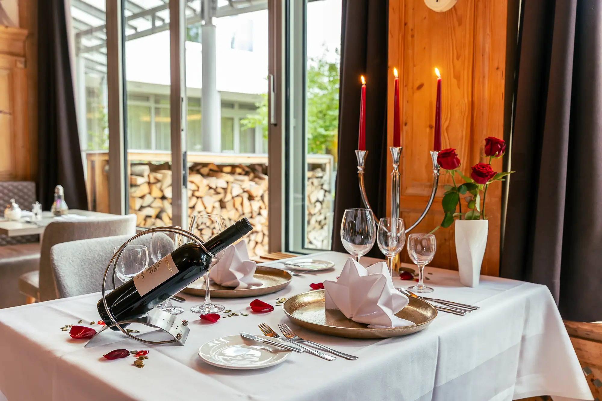 Romantisch gedeckter Esstisch mit Weinflasche, drei brennenden Kerzen und roten Rosen in einem gemütlichen Holzraum.