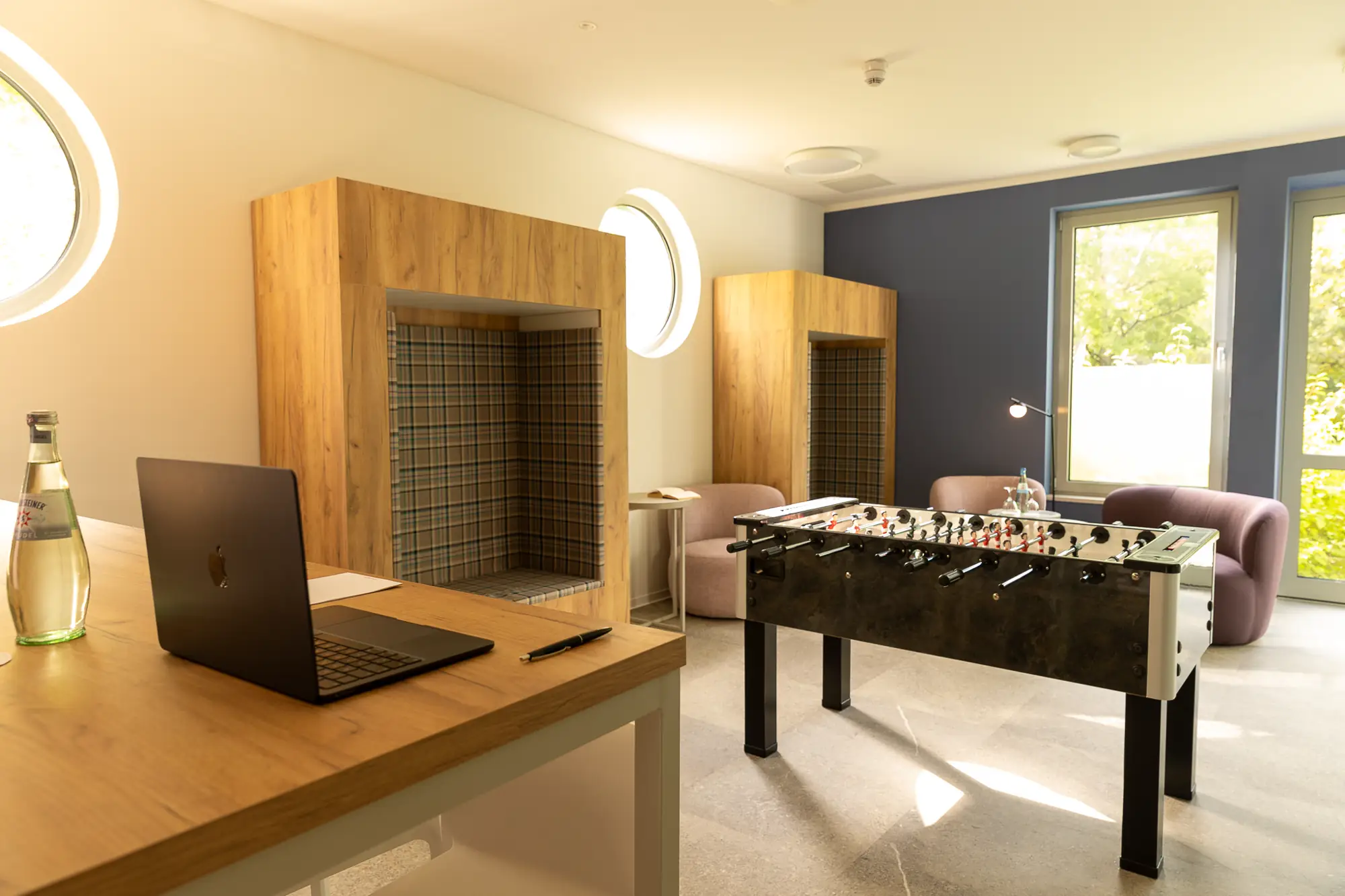Gemütlicher Aufenthaltsraum mit einem Tischkicker, Laptop auf einem Holztisch und Sitznischen mit Holzverkleidung und karierten Polstern.