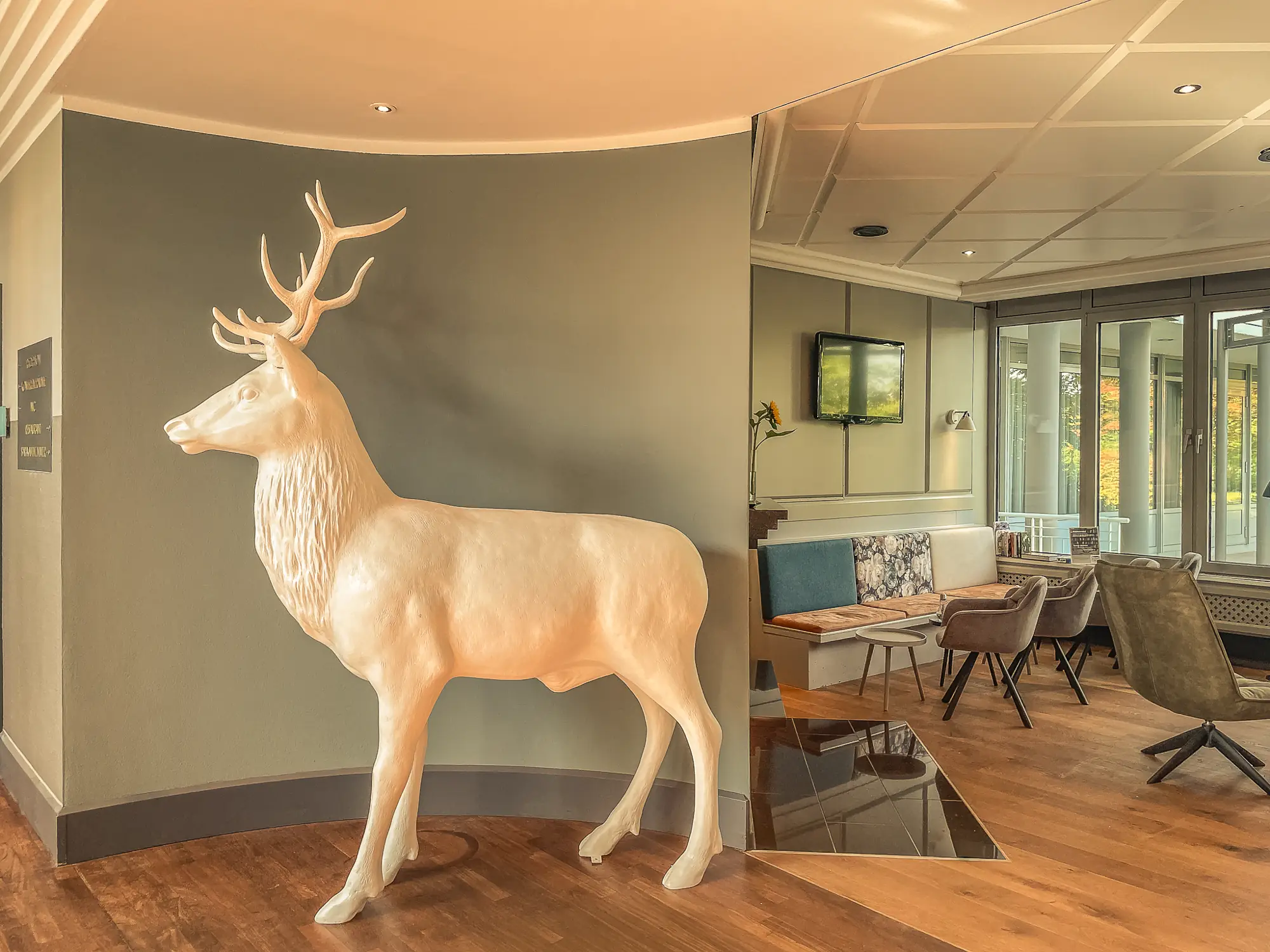 Große weiße Hirschskulptur steht vor einer grauen Wand in einem modernen Innenraum mit Holzboden und Sitzbereich.
