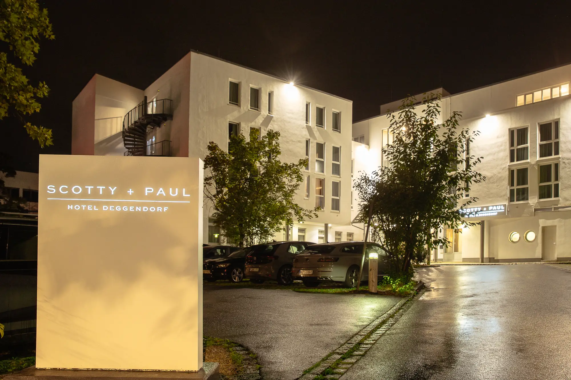 Beleuchteter Eingangsbereich des Hotels Scotty + Paul in Deggendorf bei Nacht mit geparkten Autos und Bäumen.