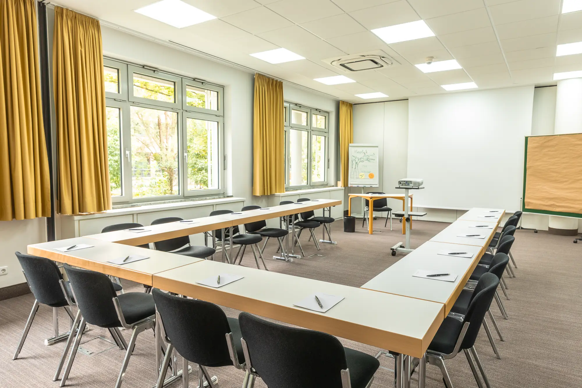 Heller Konferenzraum mit U-förmigen Tischen, schwarzen Stühlen, gelben Vorhängen und Präsentationstechnik.