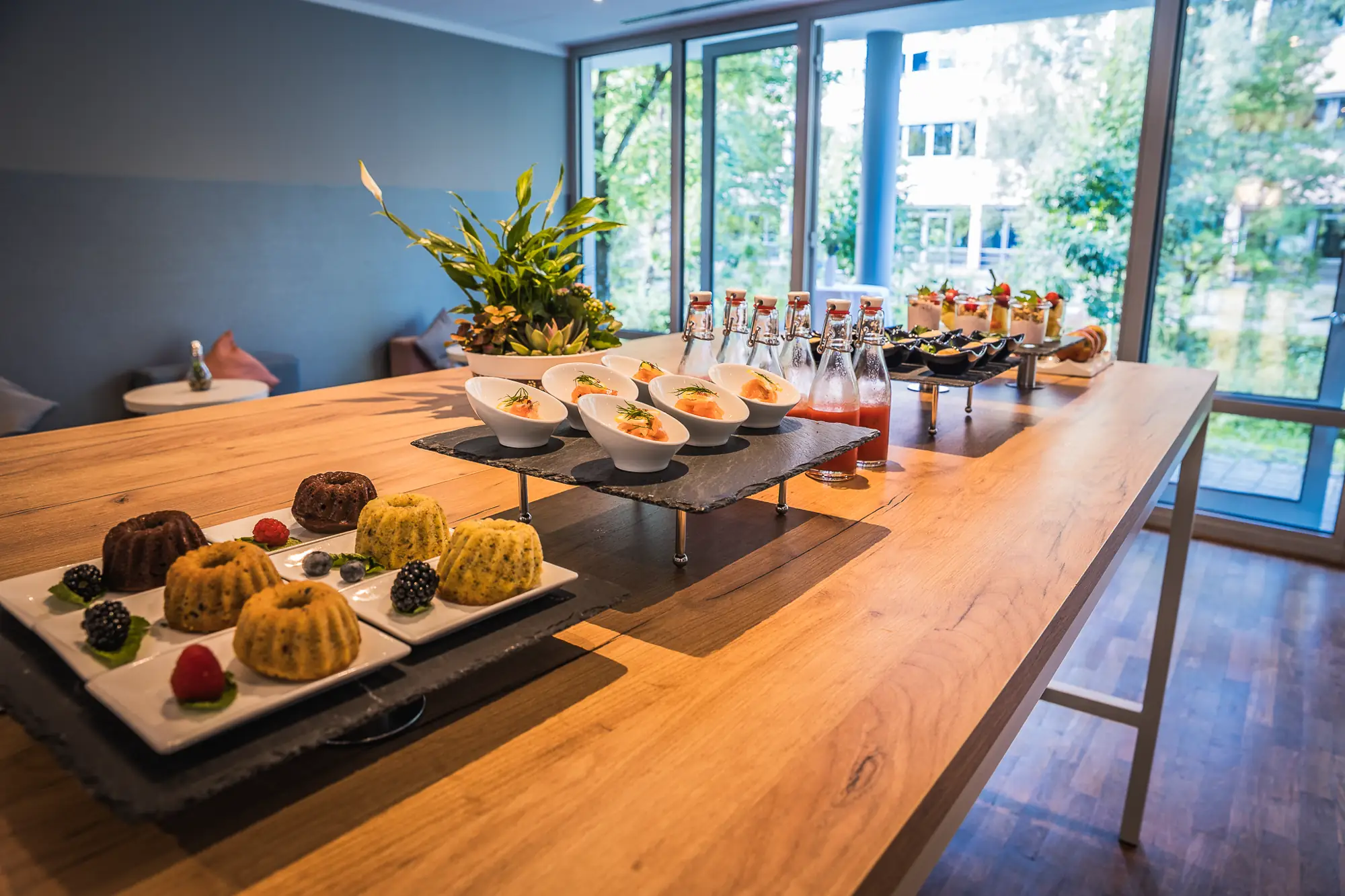 Buffet mit kleinen Kuchen, Vorspeisen in Schalen und Flaschen mit Getränken auf einem Holztisch vor großen Fenstern.