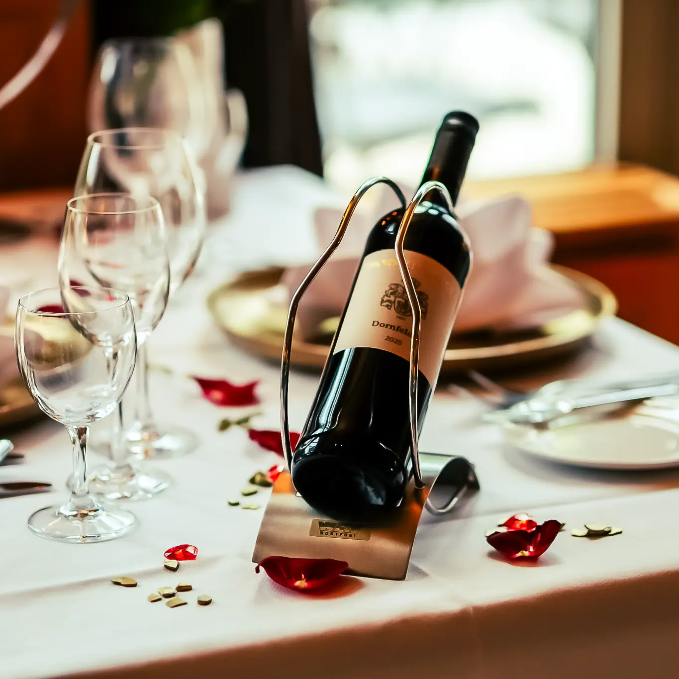 Eleganter gedeckter Tisch mit Weinflasche in Halter, Weingläsern und Rosenblättern auf weißem Tuch.