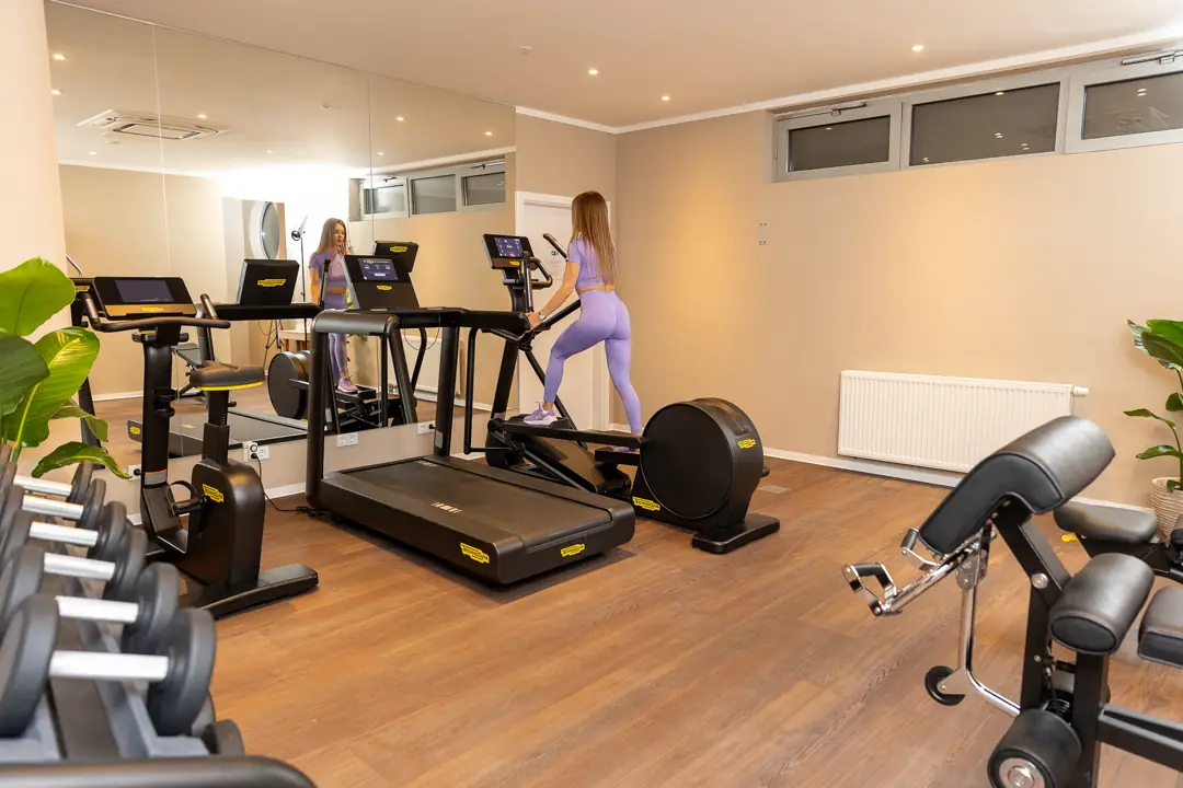 Eine Frau in lila Sportkleidung trainiert auf einem Crosstrainer in einem modernen Fitnessraum mit Holzboden, Spiegelwand und Hanteln.