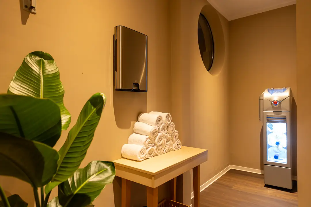 Ein Stapel weißer, gerollter Handtücher auf einem Holztisch in einem warm beleuchteten Raum mit Pflanze links und einem Wasserspender rechts.