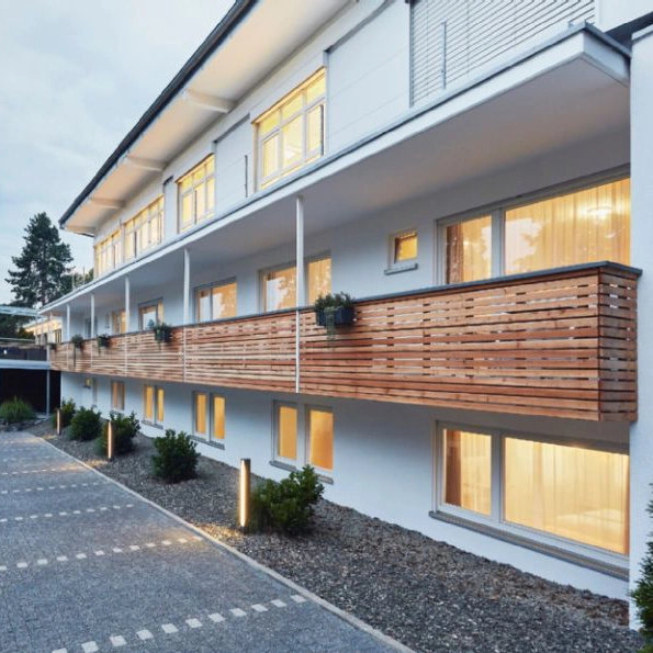 Modernes Hotel-Gebäude mit weißen Wänden, großen Fenstern und einem langen Balkon mit Holzverkleidung bei Dämmerung.