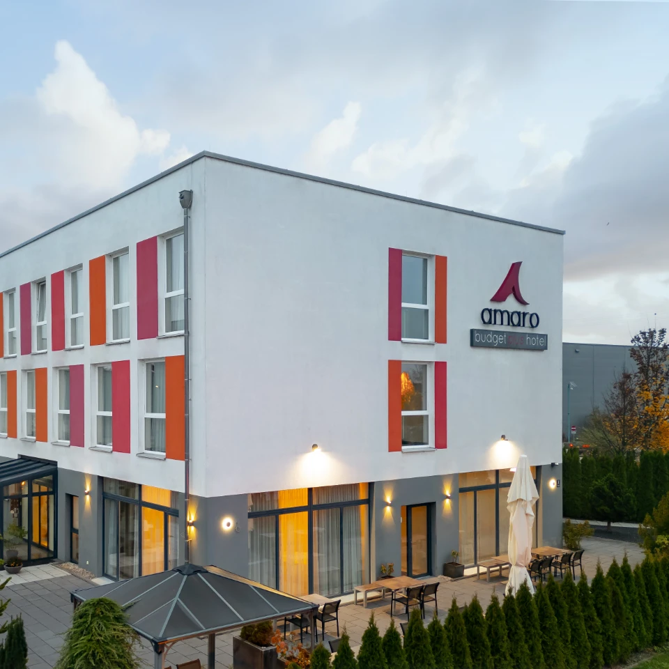 Modernes dreistöckiges Hotel mit weißer Fassade und orange-roten Fensterakzenten, beleuchtete Terrasse mit Tischen und Stühlen im Freien.