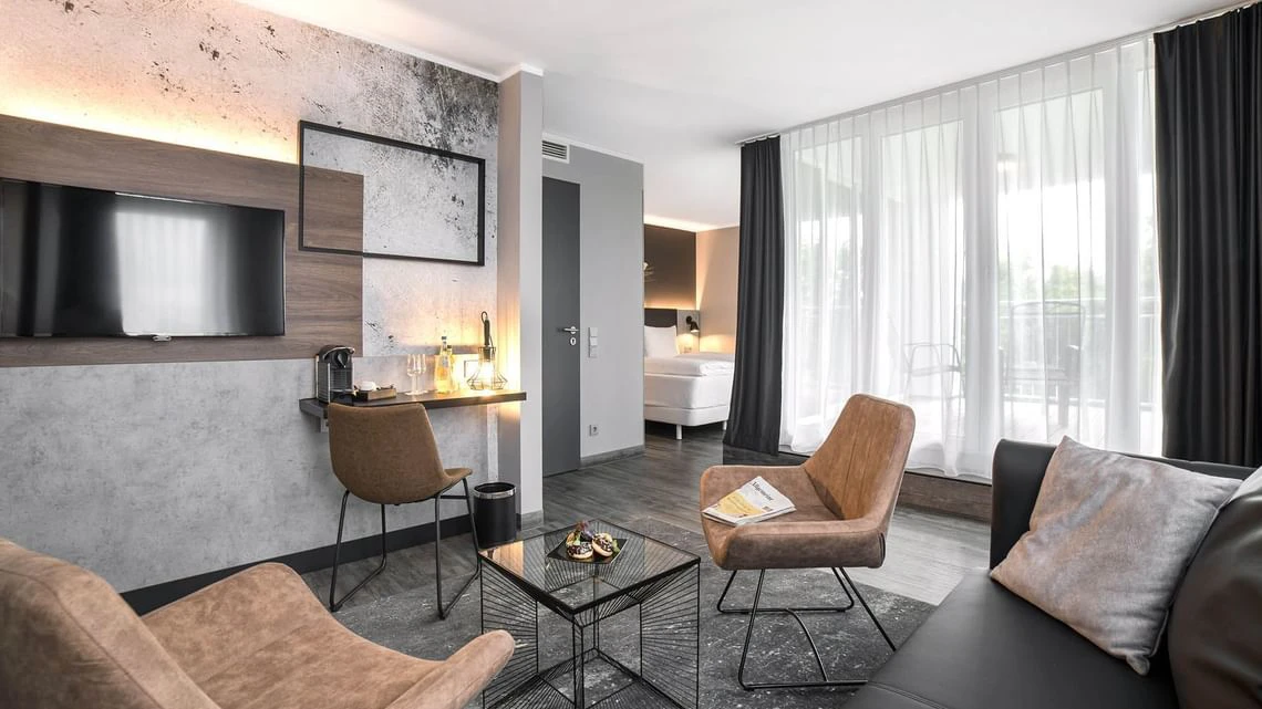 Modernes Hotelzimmer mit Sitzecke, Glas-Couchtisch, TV an der Wand und großem Fenster mit Vorhängen.
