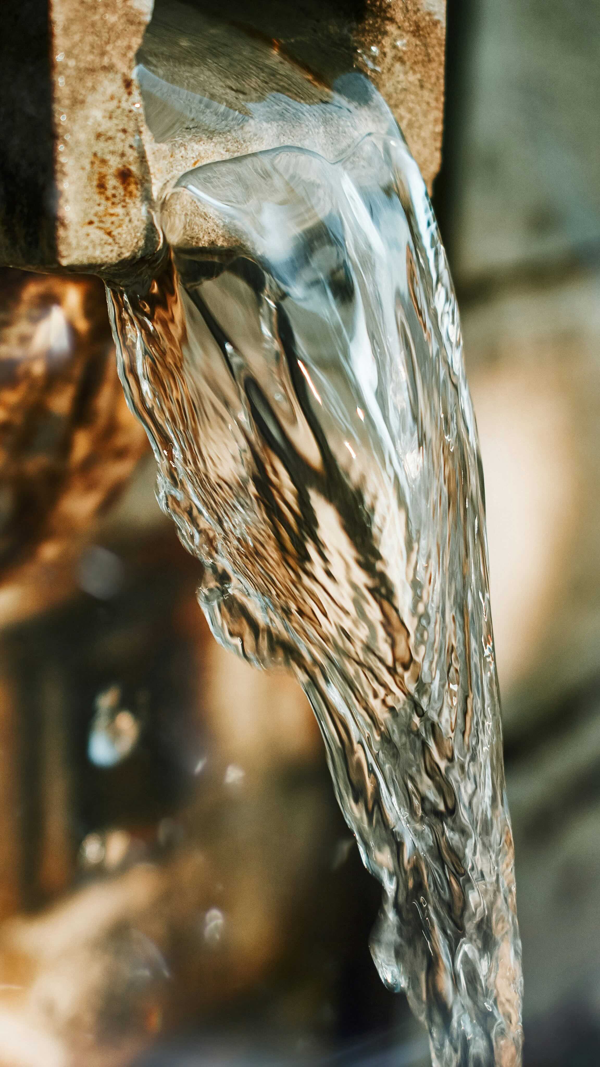 Clear water flowing smoothly from a rusty outdoor spout against a blurred background.