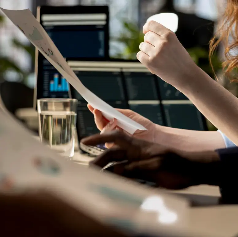 Une personne tenant un document imprimé avec un ordinateur portable affichant des graphiques en arrière-plan.