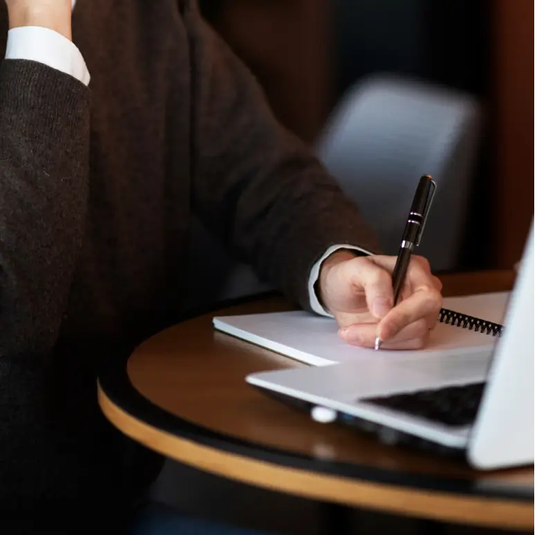 Personne écrivant dans un carnet à spirale posé sur une table à côté d'un ordinateur portable.
