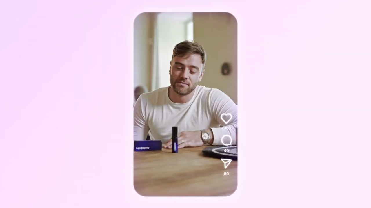Man in white shirt sitting at table, looking at a small blue bottle and box in front of him on a video screen.
