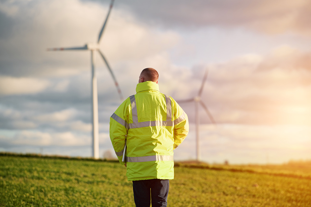 Person in high-visibility jacket stands in field facing wind turbines under cloudy sky and sunlight.