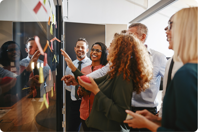 Five colleagues smiling and discussing plans using sticky notes on a glass board in an office.