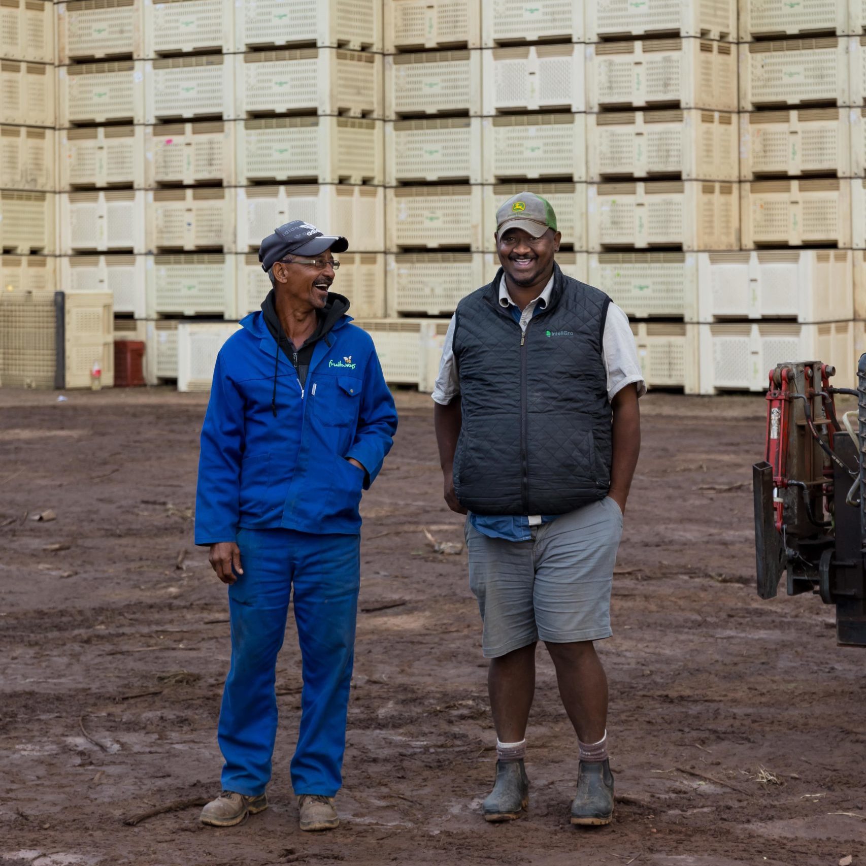 Two men standing and smiling in front of stacked crates on a muddy ground outdoors.