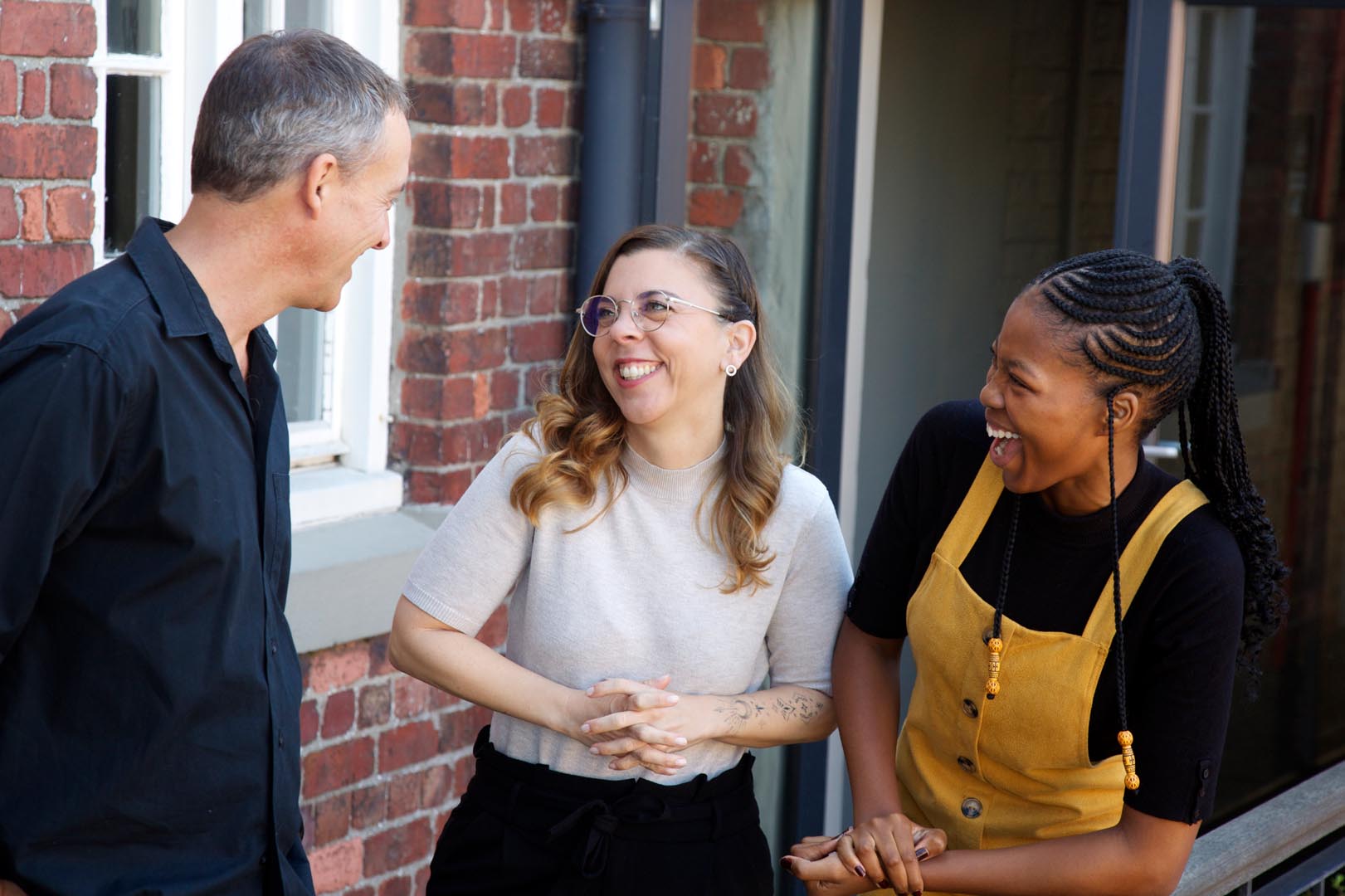 Three diverse adults laughing and chatting outside near a brick wall and window.