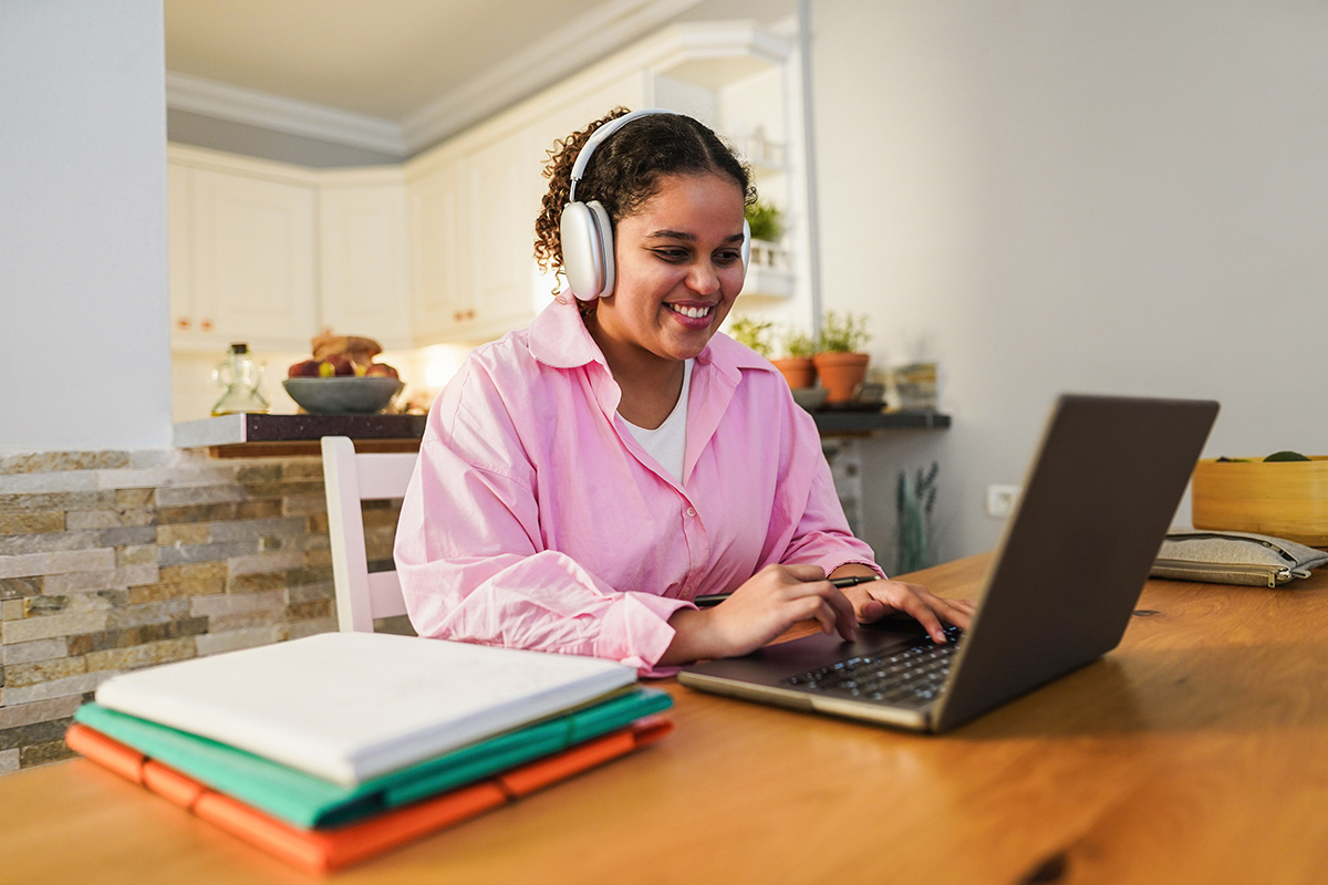 Smiling woman wearing headphones working on a laptop at a kitchen table with books nearby.