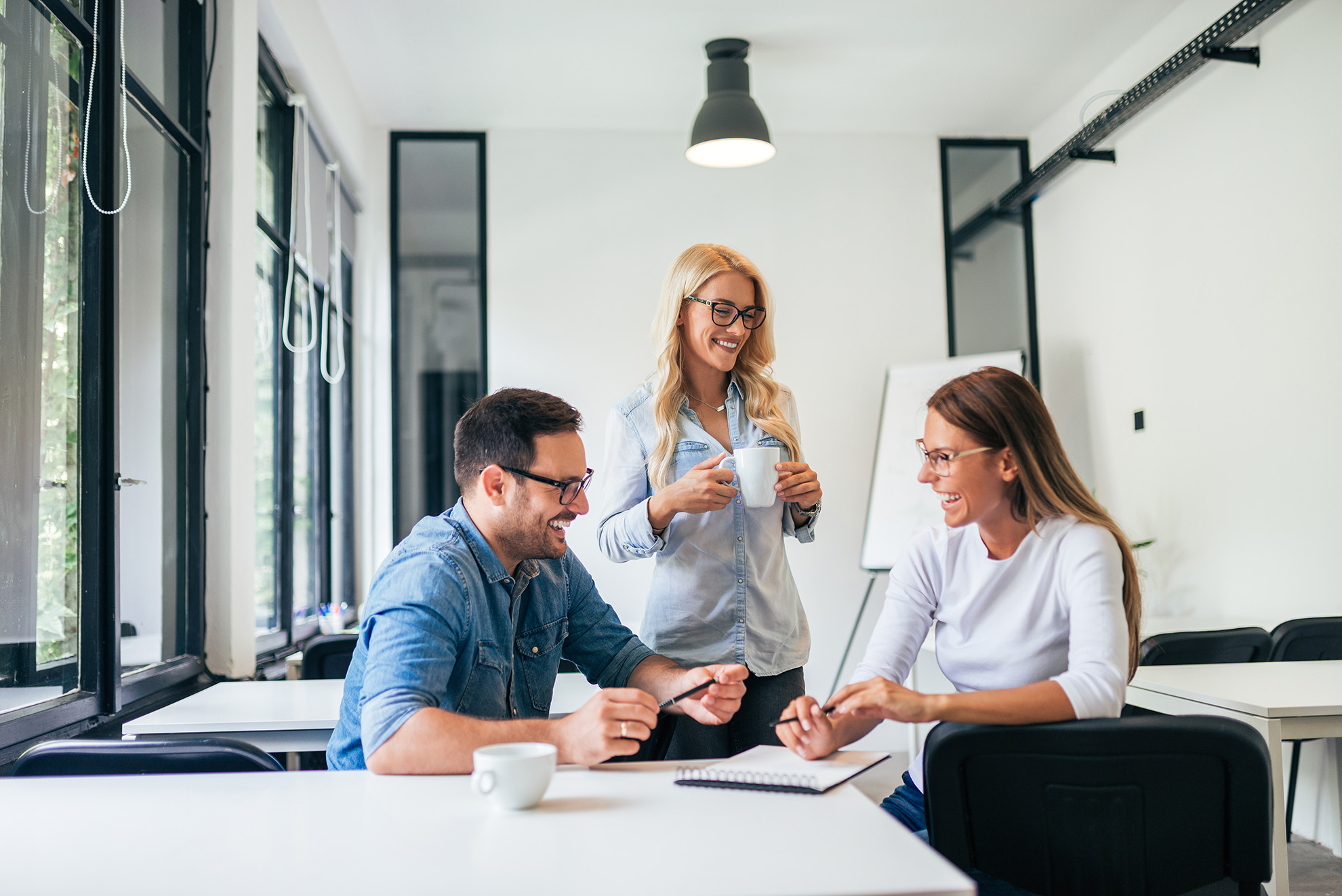 Three coworkers smiling and talking in a modern office space with coffee and a notebook.