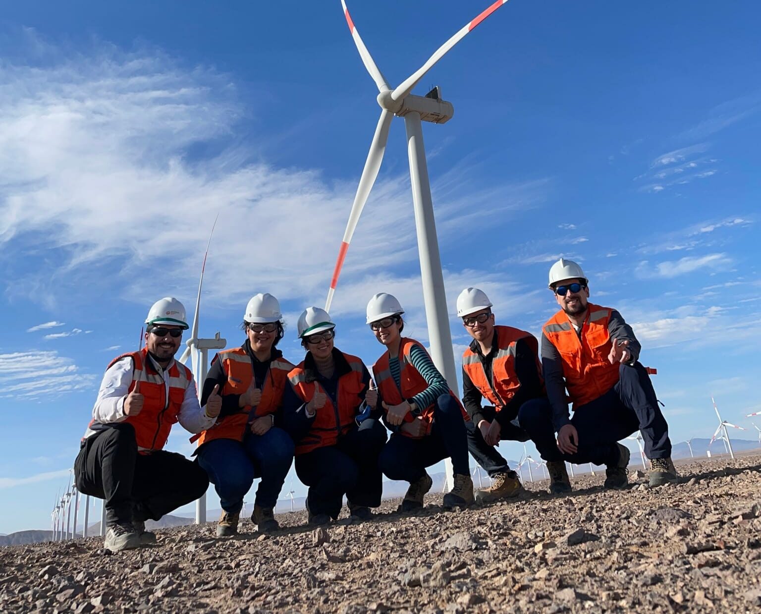 Six workers in hard hats and orange vests posing by wind turbines in a sunny desert landscape.