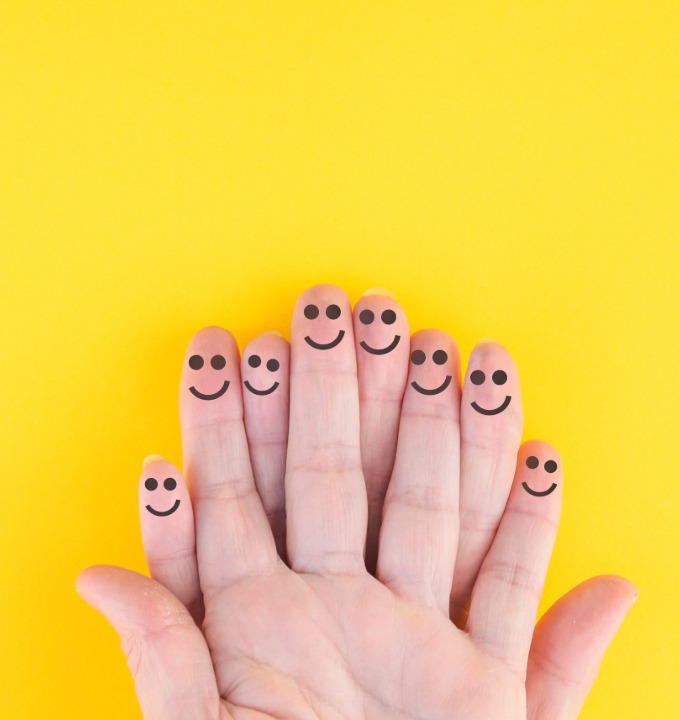 Hand with smiling faces drawn on the tips of all five fingers against a yellow background.