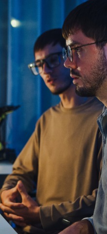 Two men wearing glasses engaged in conversation indoors with a dark blue background.