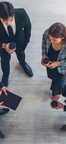 Four businesspeople standing in a circle holding coffee cups and a tablet, viewed from above.