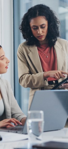 Two businesswomen, one pointing at a tablet while the other types on a laptop in an office.
