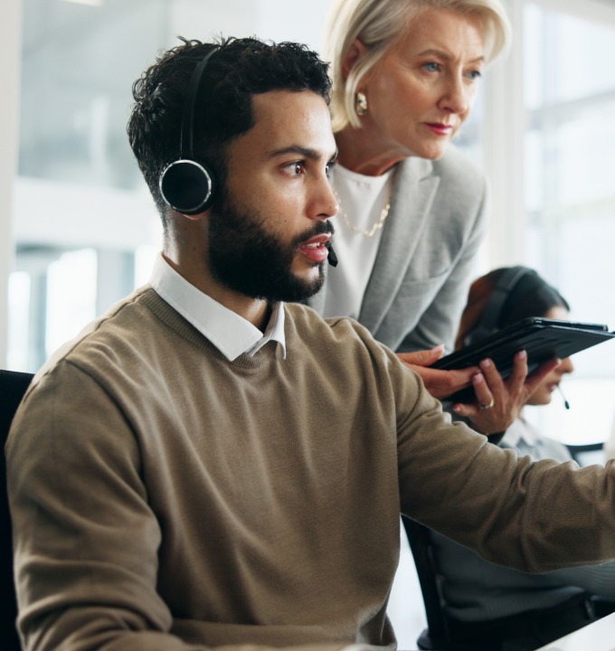 Customer service agent with headset listens while older woman reviews info on tablet nearby.