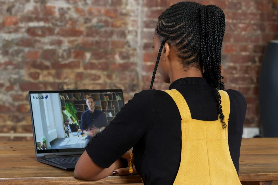 Woman with braided hair in yellow overalls sitting at a wooden table watching a man on a laptop screen.