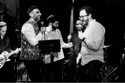 Four men singing passionately into microphones on stage with a guitarist in the background.