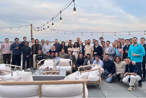 Large group of people posing together outdoors near cushioned seating and string lights at dusk.