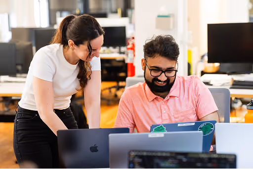 Two coworkers smiling and collaborating while looking at laptops in a modern office.