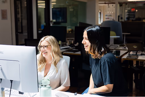 Two women smiling and working together at a computer in a modern office setting.