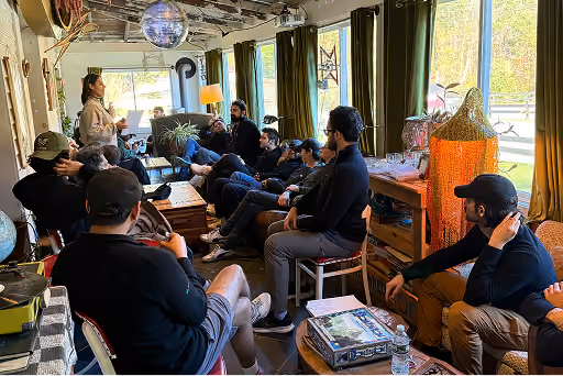 Group of people seated in a cozy room listening to a woman standing and speaking near windows with green curtains.