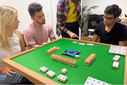Three people gathered around a table playing Mahjong, focused on the game tiles.