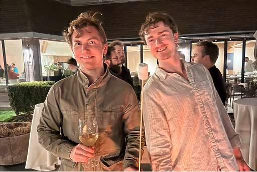 Two young men smiling at an outdoor evening gathering, one holding a glass of wine.