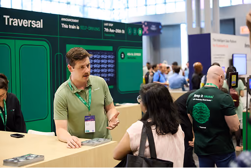 Man with a mustache explaining something to a woman at an event booth with a green backdrop.