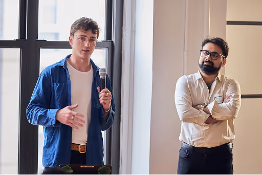 Man in blue shirt speaking into a microphone while another man in white shirt listens with arms crossed.