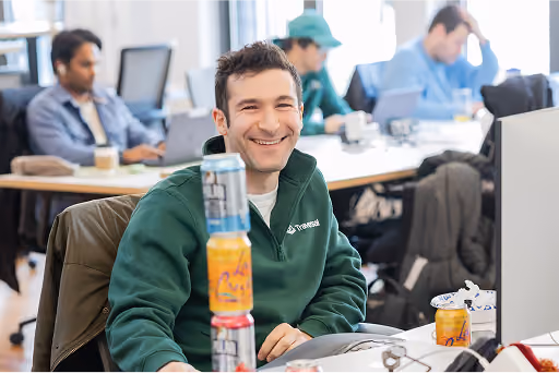 Smiling man in green jacket sitting at desk stacked with soda cans, office background with coworkers.