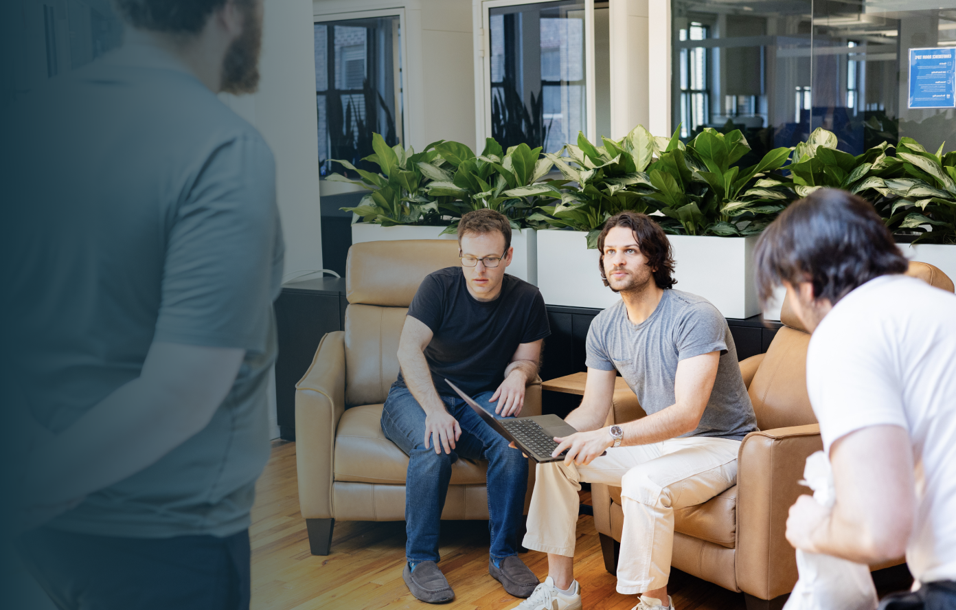 Three men in a modern office lounge, one holding a laptop, engaged in a discussion.