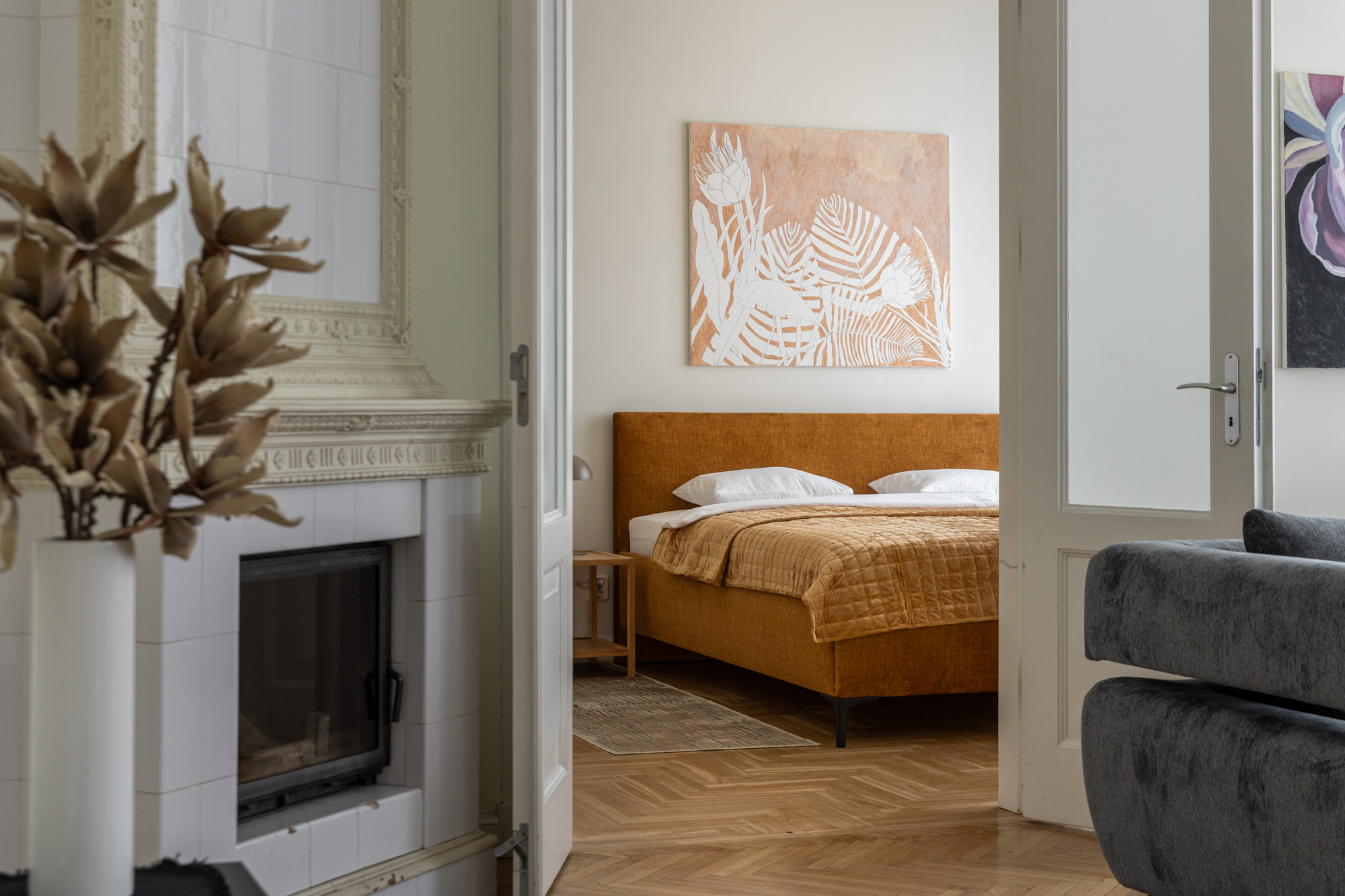 Skats uz guļamistabu ar oranžas krāsas gultu, aiz kuras ir apdrukāts rozā un balts sienas mākslas darbs, redzams arī kamīns un pelēks dīvāns.