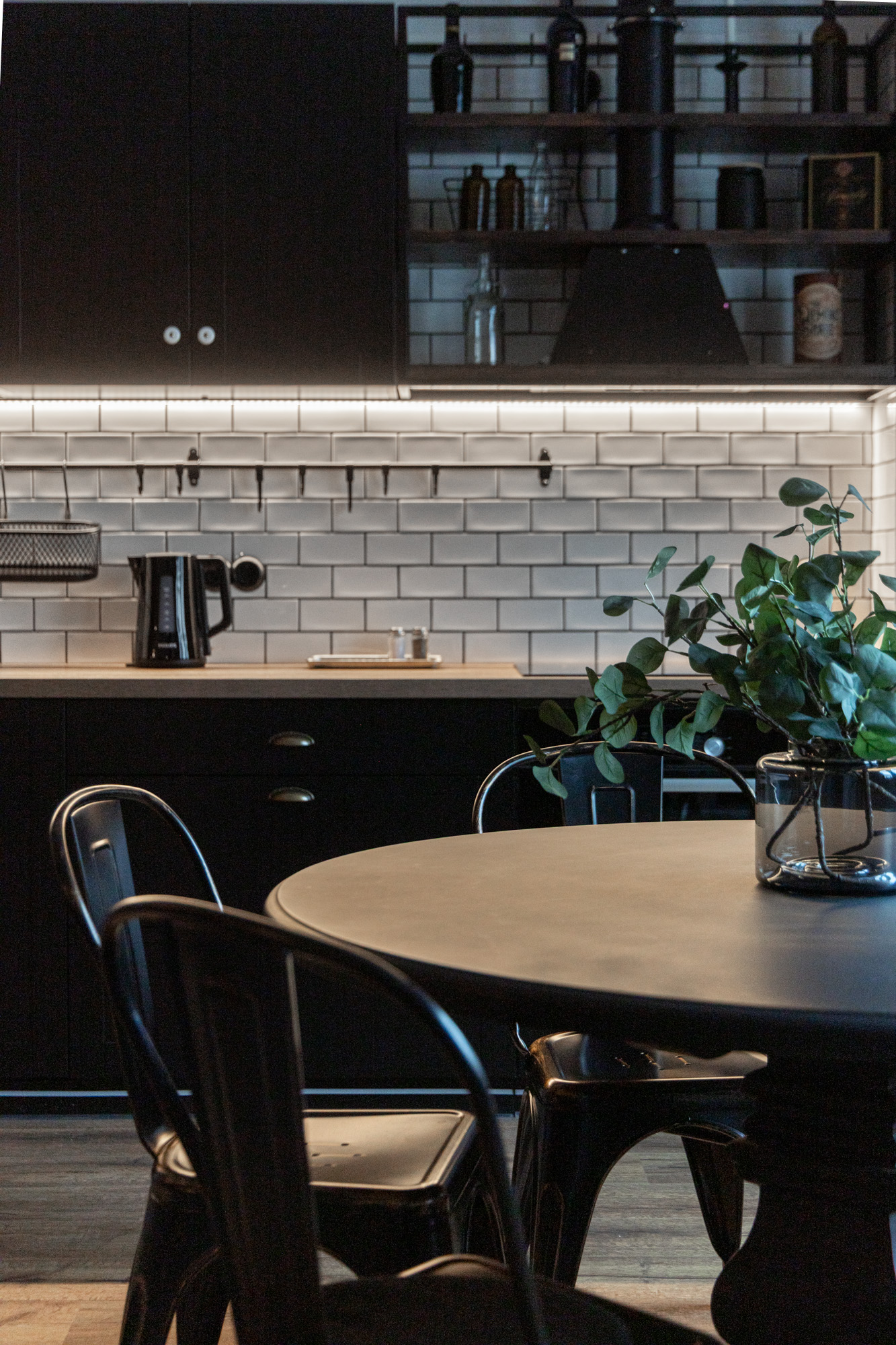 Tumša virtuves zona ar apaļu galdu, melnām metāla krēsliem un zaļas lapas stikla vāzē uz galda.