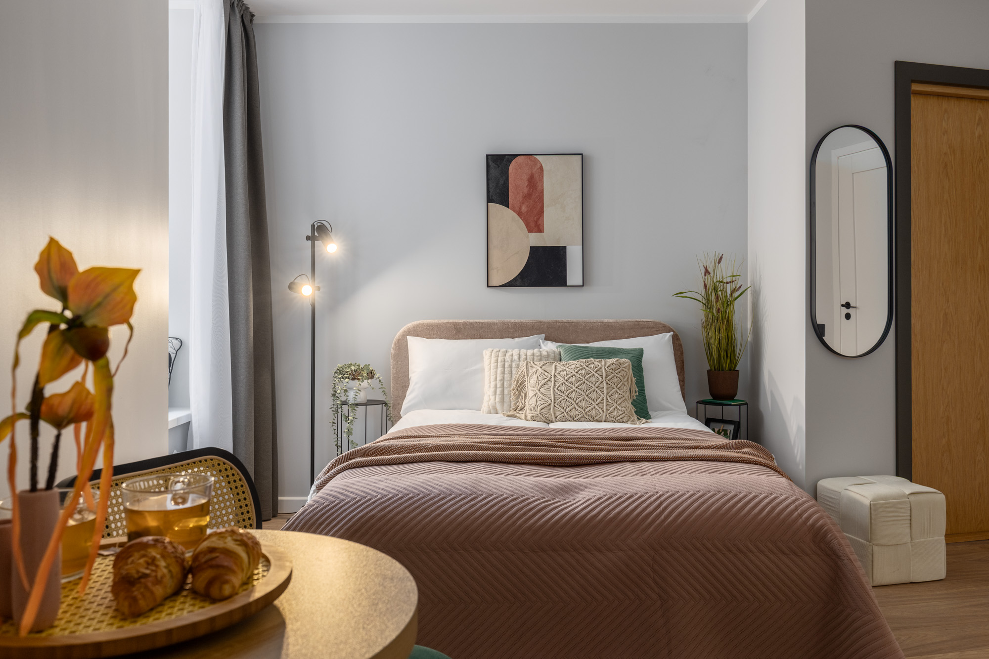 Mūsdienīga guļamistaba ar brūnu segu, dekoratīviem spilveniem uz gultas, abstraktu gleznojumu pie sienas un brokastīm ar kruasāniem uz galda.