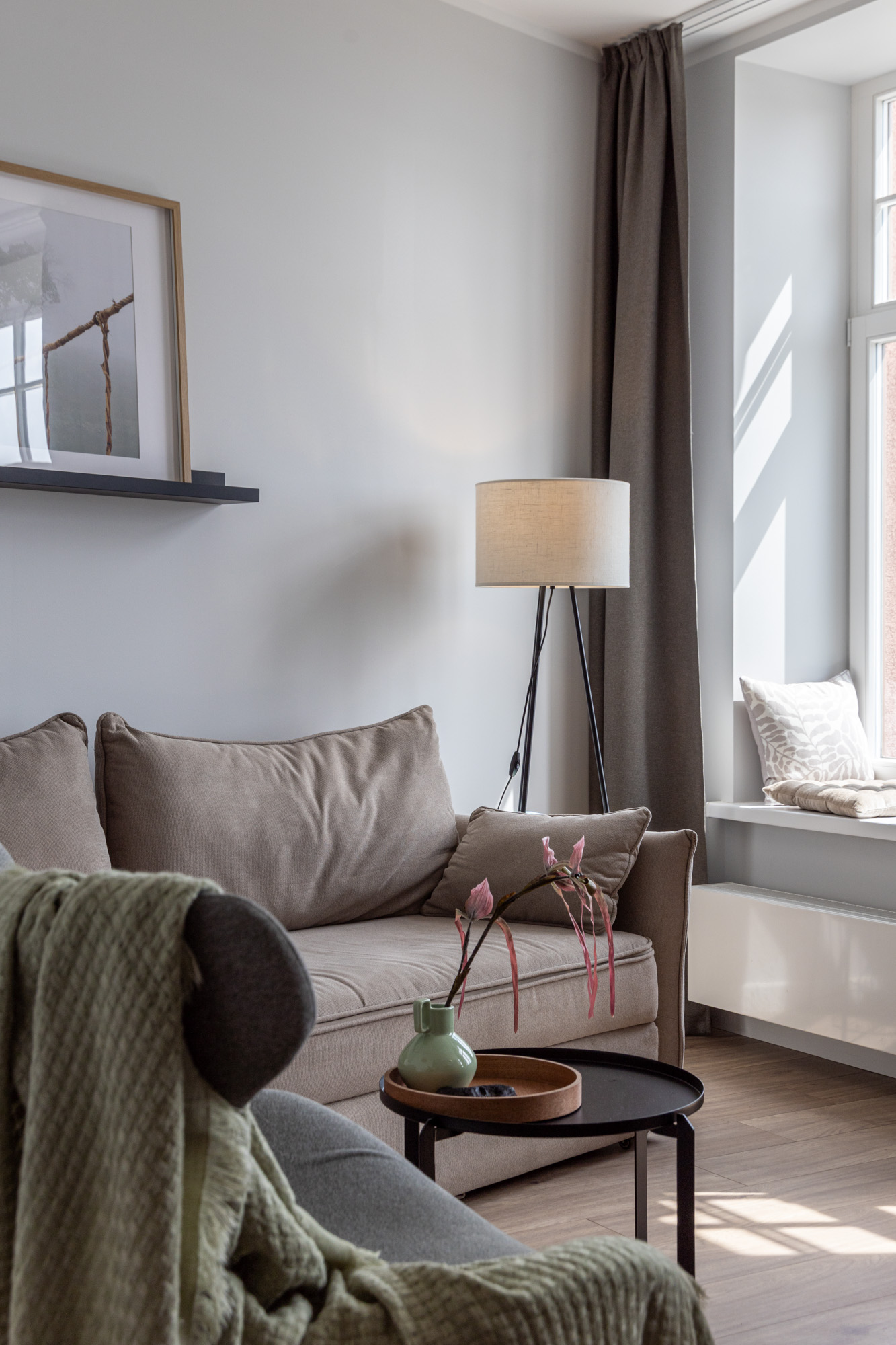 Dzīvojamā istaba ar brūnu dīvānu, stāvlampu, melnu kafijas galdiņu ar zaļu vāzi un logu ar spilvenu.