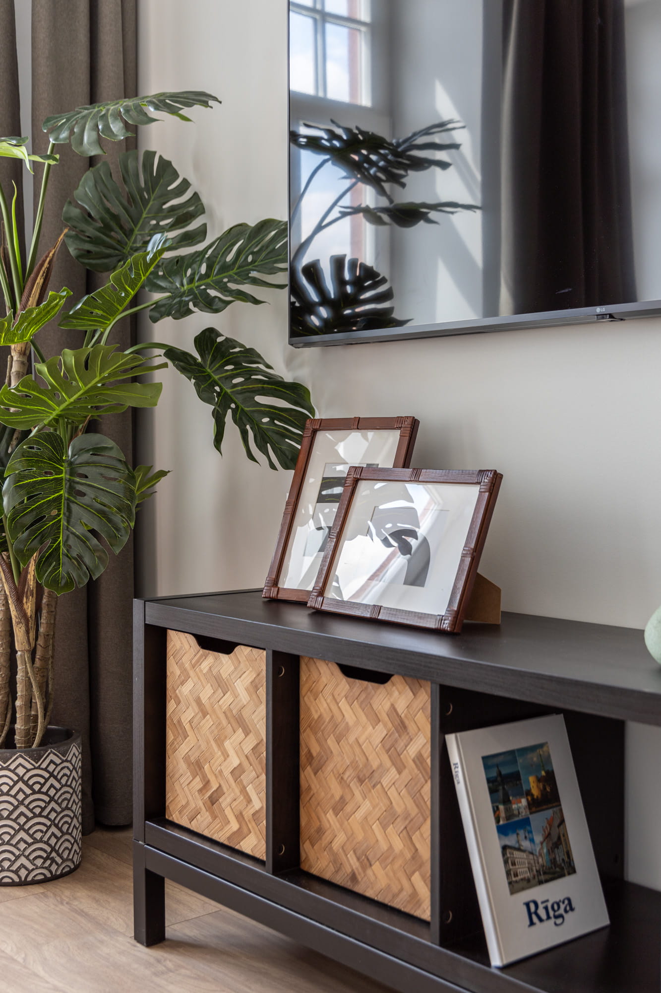 Melnas mēbeles ar divām salmu grozu kastēm, diviem rāmjiem uz galda un augiem pie sienas ar TV ekrāna atspīdumu.