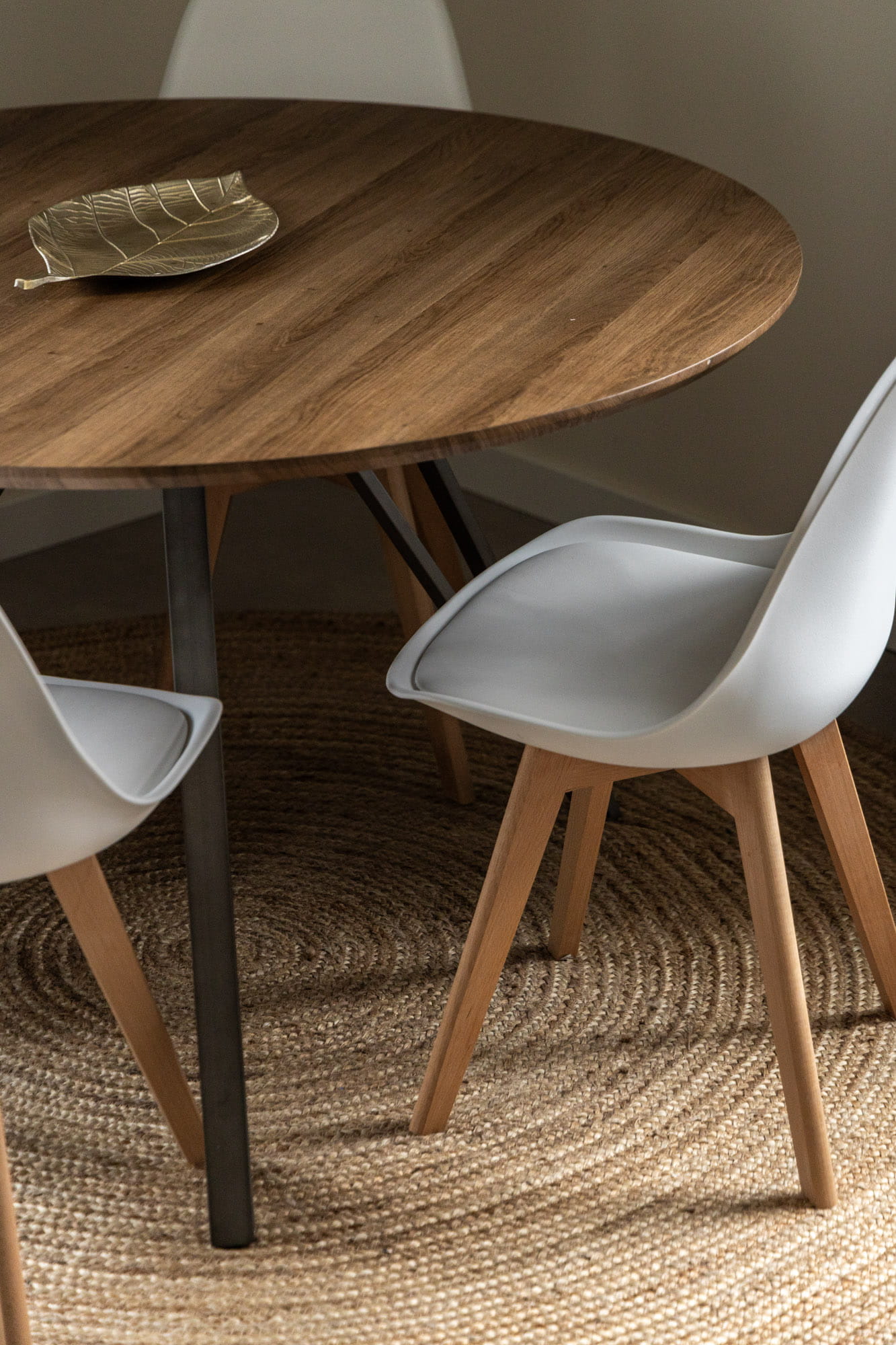 Apaļš koka galds ar divām baltām krēsliem ar koka kājām uz apaļas auztkušas paklāja.