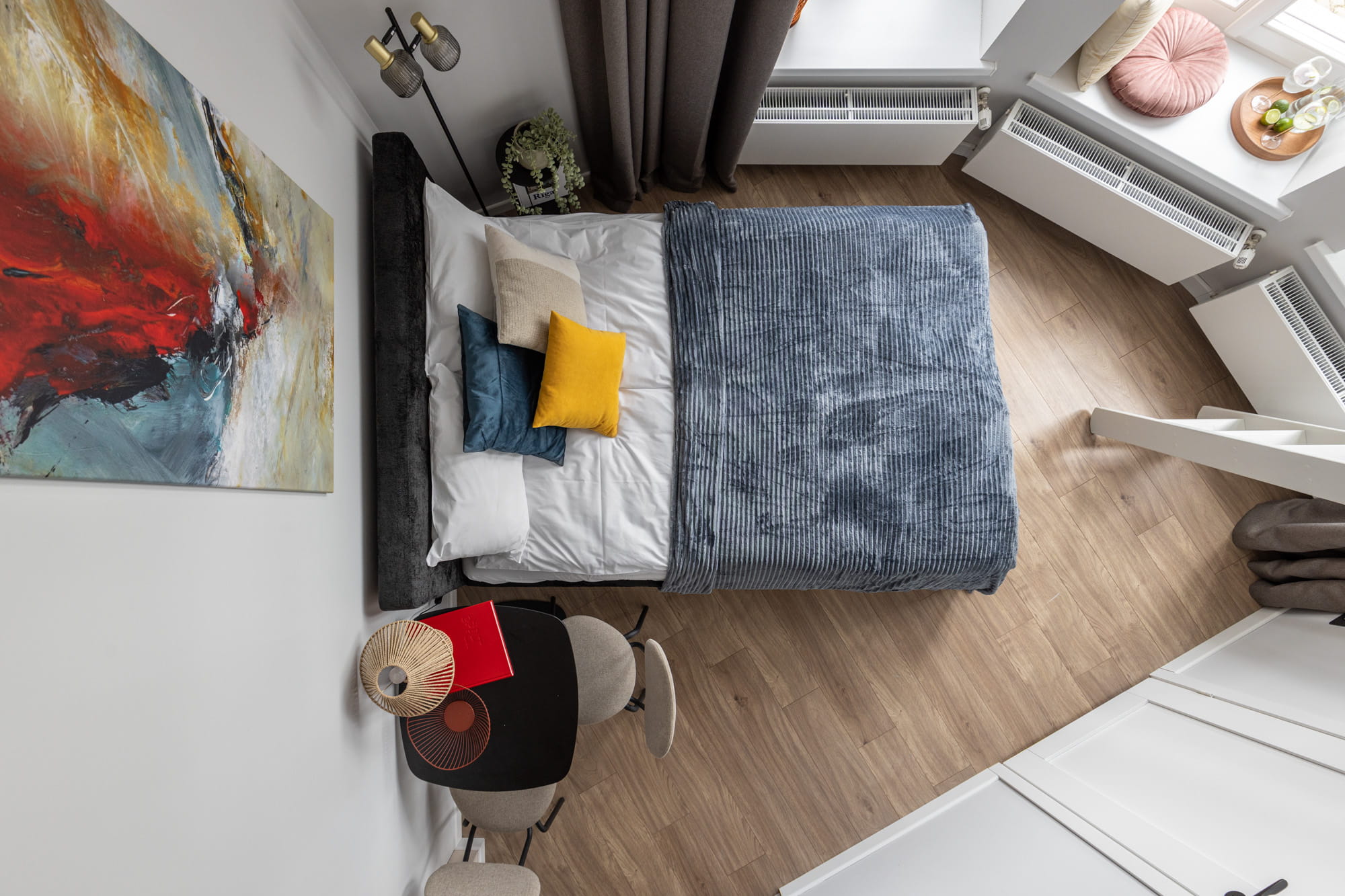 Moderna istaba ar gultu, segtu zili pelēkā vāciņā un spilveniem dažādās krāsās, mākslas gleznu uz sienas un koka grīdu.