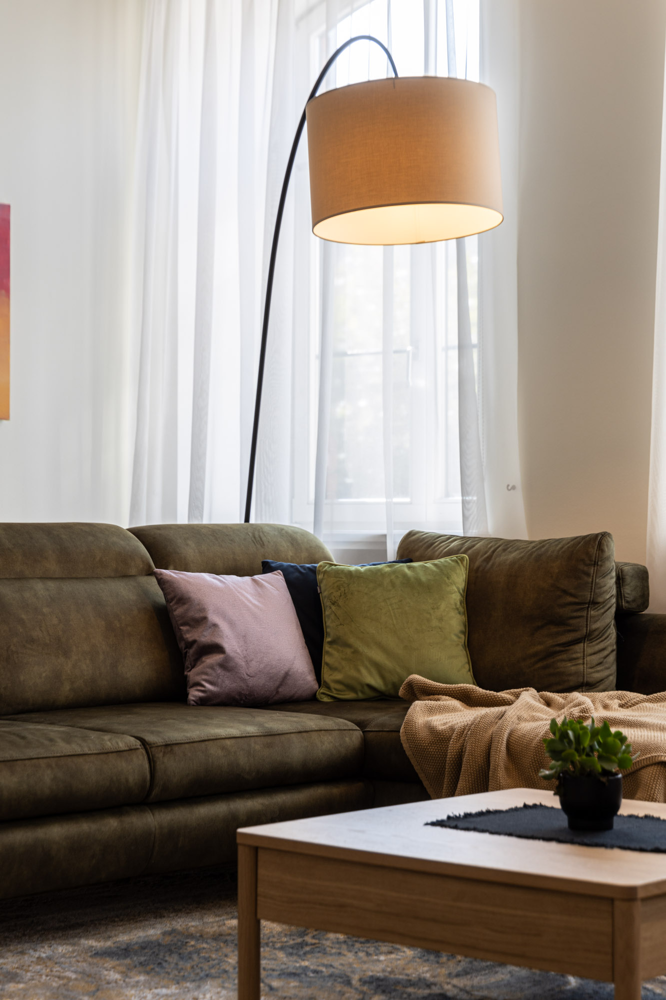 Dzīvojamās istabas stūrītis ar tumši zaļu dīvānu, trīs spilveniem, stāvlampu un kafijas galdiņu ar zaļu augu.